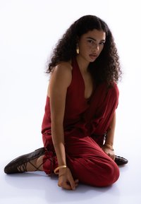 Tenue en lin rouge avec un col en V profond et une coupe ample, associée à des chaussures noires à lacets. Bracelets dorés et grandes boucles d'oreilles comme accessoires remarquables.