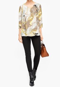 Blusa a fantasia con scollo a V, caratterizzata da vortici dorati, marroni e neri su uno sfondo chiaro, abbinata a pantaloni neri attillati e stivaletti.