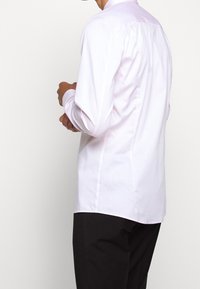 Długo rękawowa, jasnoróżowa koszula z gładkiej bawełny, z kołnierzykiem i detalami na szwie z tyłu, w zestawie z ciemnymi spodniami.