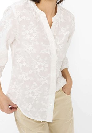 Vrouw draagt een wit blouse met knopen en bloemrijke borduur en beige broek, met één hand in de zak en de andere blouse aanpassend.