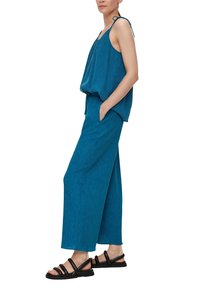 Teal mouwloze top met strikbandjes, losse pasvorm, gecombineerd met brede broek. Zwarte sandalen met bandjes. Gladde textuur met subtiel patroon.