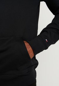 Svart sweatshirt med framficka, ribbade muddar och justerbar huva. Har en liten logodetalj på ärmen. Mjukt, texturerat tyg.