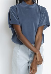 Blå kortärmad topp med hög krage, mjuk textur och avslappnad passform; bärs med ljusa denimbyxor.