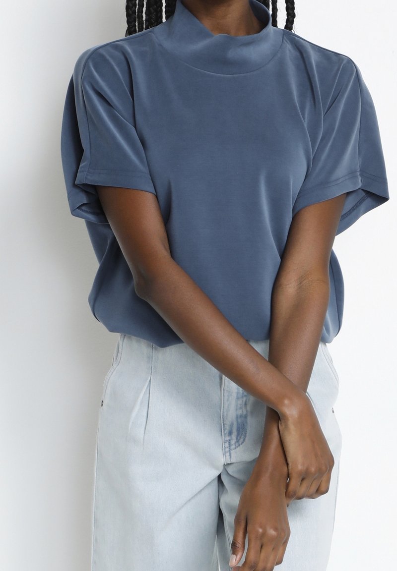 Blå kortärmad topp med hög krage, mjuk textur och avslappnad passform; bärs med ljusa denimbyxor.