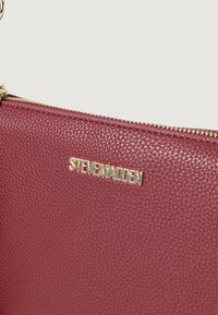 Burgundi textúrájú bőrtáska arany színű "STEVE MADDEN" logóval és cipzár részletekkel. Kompakt formatervezés sima felülettel.