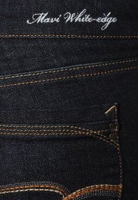 Vaqueros de denim azul oscuro con costuras naranjas, que cuentan con un pequeño bolsillo y un logotipo de marca bordado en blanco a lo largo de la cinturilla.