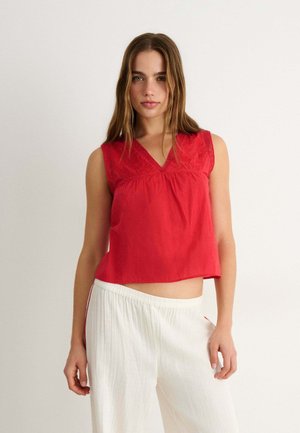Femme aux longs cheveux bruns portant un haut rouge sans manches à col en V et un pantalon blanc ample, debout devant un fond clair uni.
