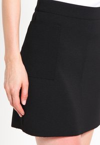 Svart kjol i texturerad tyg med sidoficka, rak skärning och rå kant. Nära vy med handen placerad vid midjan.