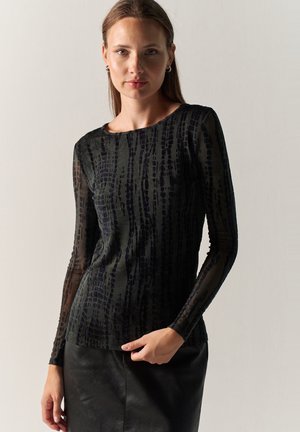 Mujer con cabello castaño liso con una blusa negra transparente con estampado y mangas largas y falda negra, de pie contra un fondo claro y liso.
