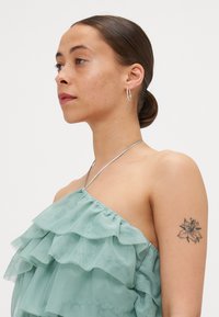 Lys myntegrønn kjole med lagdelte volanger i gjennomsiktig stoff, med halterneck og et sølvhalskjede. Flekket hud med en blomstertatovering.