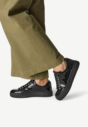 Sneakers din piele patent neagră cu o talpă groasă neagră, vârful rotund și ochiuri argintii, asortați cu pantaloni largi de culoare verde oliv.