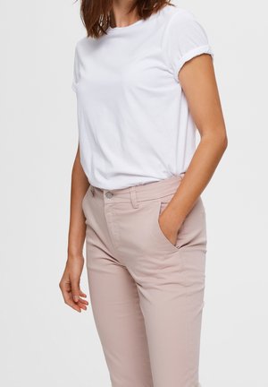 Femme portant un t-shirt blanc uni à manches courtes rentré dans un pantalon beige clair ajusté, debout avec une main dans la poche.