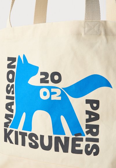 Grafică albastră stilizată a unei vulpi cu „20 02” în interior, înconjurată de text negru cu inscripția „Maison Kitsuné Paris” pe o geantă din pânză bej.