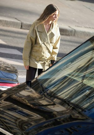 Joven mujer con una chaqueta beige de gran tamaño, de pie en la calle cerca de coches, mirando hacia un lado a la luz del día.