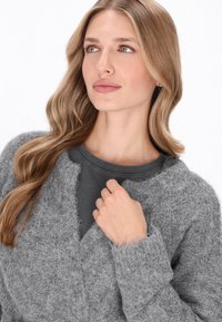 Grauer Strickcardigan mit strukturiertem Finish, mit V-Ausschnitt und lockerer Passform, über einem dunkelgrauen, gerippten Shirt getragen.