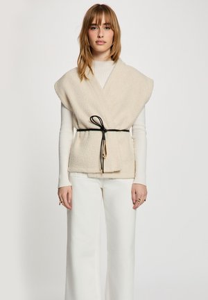 Femme portant un gilet sans manches texturé crème noué avec une ceinture noire sur un haut blanc à manches longues côtelé et un pantalon blanc, debout devant un fond uni.
