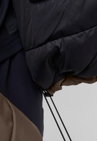 Giacca a vento imbottita in nylon blu scuro con dettagli marroni. Le maniche presentano polsini elasticizzati e un dettaglio con cordino per la regolazione.