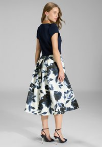 Marineblaues Oberteil mit kurzen Ärmeln, kombiniert mit einem knielangen Rock in Weiß, der große Blumenmuster in Blau und Schwarz aufweist, getragen mit Riemchen-High Heels.