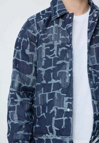 Veste en denim présentant une base bleu foncé avec un motif abstrait bleu clair. Le design comprend un col classique et une fermeture à boutons-pression.