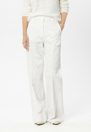 Frau trägt hochtaillierte, weit geschnittene weiße Hosen mit dezenten Pailletten, kombiniert mit einem strukturierten weißen Pullover, die Hände in den Taschen.