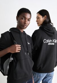 Schwarze Hoodies, präsentiert an zwei Modellen, mit einer vorderen Tasche, Kordeln und dem Schriftzug "Calvin Klein JEANS" auf dem Rücken. Glatte Stofftextur.
