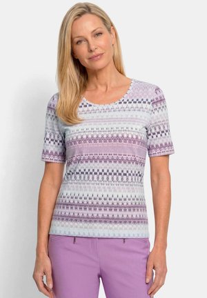 Femme aux cheveux blonds portant un haut à manches courtes à motif violet clair et blanc et un pantalon uni violet clair, debout devant un fond uni.