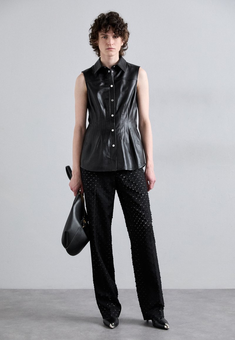 Haut sans manches en cuir noir avec des coutures structurées ; col montant. Pantalon noir avec un motif circulaire texturé ; chaussures noires à bout pointu ; tenant un sac noir.