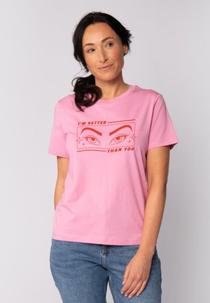 Femme aux cheveux bruns attachés en queue de cheval portant un t-shirt rose avec des yeux graphiques rouges et le texte « JE SUIS MEILLEURE QUE TOI », associé à un jean bleu.