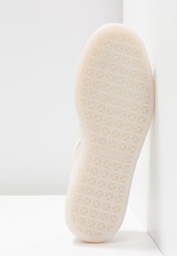 Semelle d'une basket blanche montrant un motif géométrique texturé pour l'adhérence, positionnée verticalement contre un angle de mur et de sol blancs.