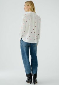White shirt with red lip patterns, long sleeves, soft fabric, and a straight cut. Paired with light blue relaxed-fit jeans and black ankle boots.