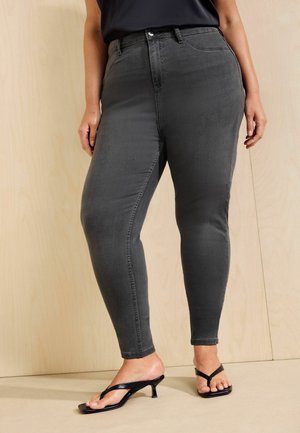 Person trägt dunkelgraue Skinny-Jeans und schwarze Sandalen mit Absatz und steht vor einer hellen Holzwand.