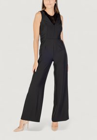 Mono negro con un diseño de escote en V y detalles de encaje transparente, silueta de pierna ancha y textura suave. Llevado con tacones nude.