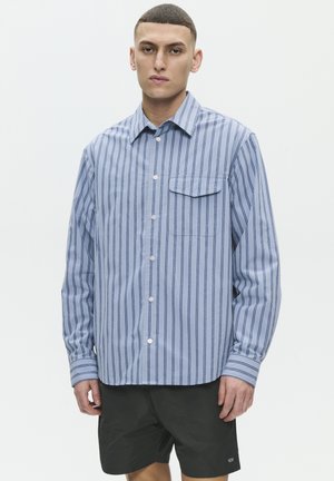 Jeune homme portant une chemise bleu clair à boutons avec des rayures verticales bleu foncé et un short noir, debout devant un fond blanc uni.