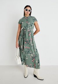 Robe maxi florale verte, avec un col, des manches courtes, une ceinture à nouer et un motif de fleurs roses. Associée à des bottes de cowboy blanches et un sac à main.