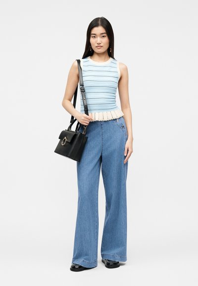 Νεαρή γυναίκα με μακριά μαύρα μαλλιά, που φοράει μπλε ριγέ αμάνικο τοπ, τζιν παντελόνι με φαρδιά μπατζάκια, μαύρα παπούτσια και κρατάει μια μαύρη τσάντα.
