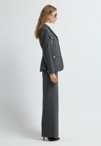 Traje gris a medida con una chaqueta de doble botonadura que presenta bolsillos con solapa y botones plateados, combinado con pantalones de pierna recta y tacones altos.