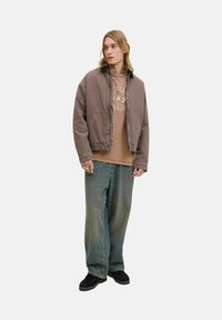 Blanding af bomuld, brun, cropped jakke med lynlås, sidelommer; lagd ovenpå en beige grafisk t-shirt og løse blå denim jeans. Sorte sko.