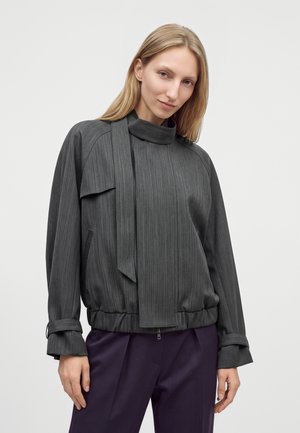 Femme aux cheveux blonds et raides porte une veste gris foncé à col haut avec des manches longues et un pantalon violet foncé sur un fond uni.