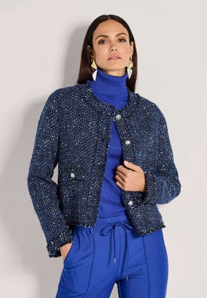 Gewebte marineblaue Jacke mit silbernen Knopfverschlüssen, strukturiertem Stoff und taillierter Passform. Kombiniert mit einem blauen Rollkragenpullover und marineblauen Hosen.