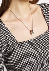 Silberkette mit einem runden Anhänger, getragen von einer Person in einem schwarz-weißen Hahnentritt-Muster-Oberteil mit quadratischem Ausschnitt.