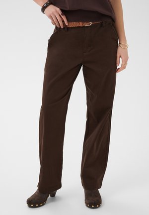 Femme portant un pantalon large marron foncé avec une ceinture tressée, un haut marron foncé et des chaussures à talons marron foncé cloutées, debout, une main sur la hanche.
