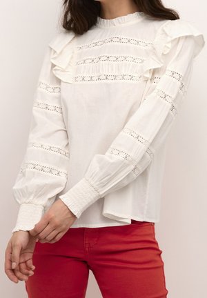 Femme portant un chemisier blanc à manches longues avec des détails en dentelle et volants, et un pantalon rouge, ajustant sa manche gauche avec sa main droite.