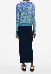 Neulottu neule sinivihreällä kuviolla, resorireuna, yhdistetty istuvaan tummansiniseen hameeseen ja mustiin kenkiin, joissa on valkoisia kukka-aksentteja.