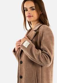 Abrigo de lana beige con tejido texturizado, cuello con muesca y botones negros. Cuenta con bolsillos laterales y una etiqueta en la manga.