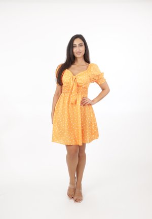 Frau steht mit der Hand in der Hüfte, trägt ein orangefarbenes Kleid mit Punkten und Puffärmeln sowie nackte, offene Schuhe vor einem weißen Hintergrund.