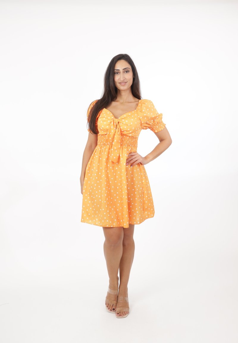 Mujer de pie con la mano en la cadera, vistiendo un vestido naranja con lunares, mangas abullonadas y tacones abiertos color nude, sobre un fondo blanco.