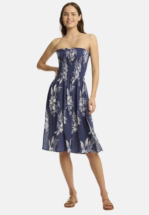 Strapless marineblauwe jurk met witte bloemenprint, smocklichaam, vloeiende rok en enkellange stijl. Geaccentueerd met platte sandalen.