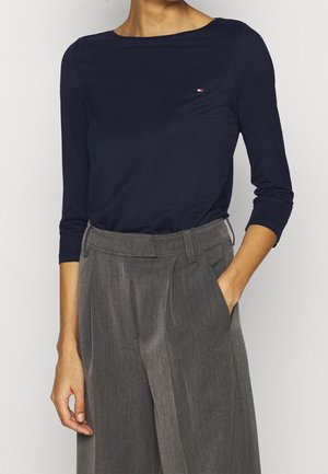 Personne portant un haut bleu marine à manches longues et un pantalon large gris taille haute, une main dans la poche.