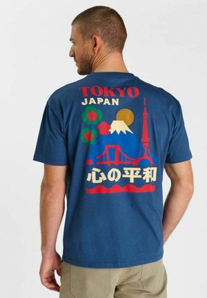 Uomo con camicia blu con disegno Tokyo Giappone che mostra il Monte Fuji, ponte rosso, torre, fiori, sole e caratteri giapponesi sulla schiena.