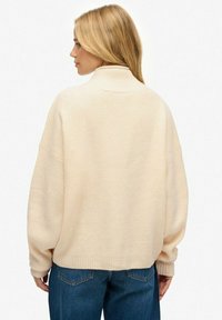 Femme aux longs cheveux blonds portant un pull à col roulé crème et un jean bleu, vue de dos sur un fond blanc.